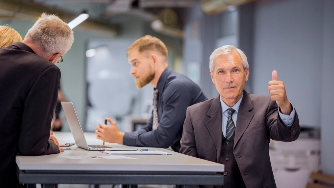 senior-businessman-showing-thumb-up-sign-sitting-front-his-team-discussing-office